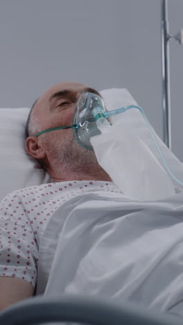 Senior-man-in-oxygen-mask-lies-in-bed-in-hospital-ward.-Old-patient-during-artificial-lung-ventilation.-Modern-emergency-room-in-clinic.-Intensive-care-coronavirus-department-in-medical-facility.-Vertical-shot