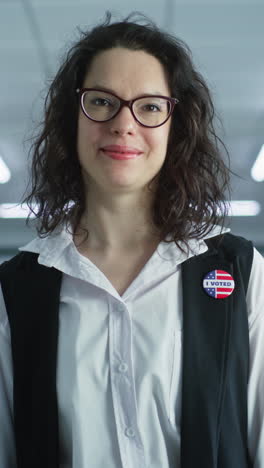 Portrait-of-Caucasian-woman,-United-States-of-America-elections-voter.-Woman-in-glasses-stands-in-polling-station,-poses,-smiles,-looks-at-camera.-Background-with-voting-booths.-Concept-of-civic-duty.