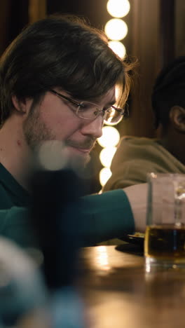 Vertical-shot-of-man-in-glasses-sits-at-bar-counter,-he-drinks,-eats-and-talks-with-African-American-friend.-People-enjoying-their-weekend-evening-in-sports-pub.-Friends-spending-time-together-in-bar.-Close-up.