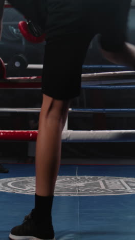 Vertical-shot-Teenager-sits-near-boxing-ring-and-puts-on-boxing-gloves.-Young-boxer-comes-in-boxing-ring-and-starts-practice-punches-with-African-American-trainer.-Athlete-prepares-to-competition-in-boxing-gym.