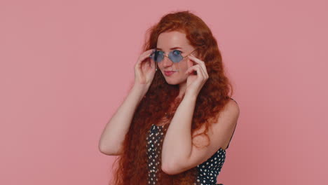 Seductive-cheerful-ginger-stylish-girl-in-dress-wearing-sunglasses,-charming-smile-on-pink-wall