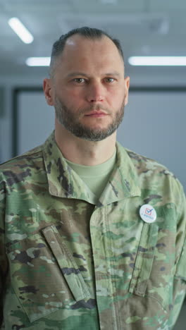 Portrait-of-male-soldier,-United-States-of-America-elections-voter.-Man-in-camouflage-uniform-stands-in-a-modern-polling-station-and-looks-at-camera.-Background-with-voting-booths.-Civic-duty-concept.