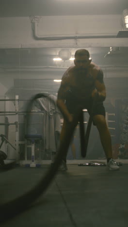 Professional-male-boxer-does-cardio-or-endurance-workout-before-championship-fight.-Athletic-man-exercises-with-battle-ropes-in-dark-boxing-gym.-Vertical-shot