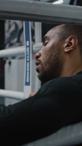 Tired-and-exhausted-African-American-boxer-sits-in-boxing-ring-corner.-Fighter-with-beads-of-sweat-on-face-rests-after-workout.-Athlete-prepares-to-tournament-or-competition-in-boxing-gym.-Vertical-shot