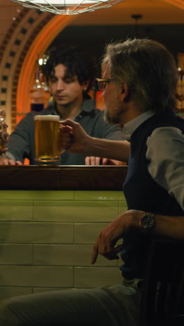 Vertical-shot-of-Young-bartender-gives-glasses-of-beer-to-mature-male-friends.-Two-men-talk-and-drink-beer-sitting-at-the-bar-counter-in-stylish-pub-at-night.-Meeting-of-old-friends.-Concept-of-lifestyle-and-leisure.