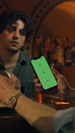 Vertical-shot-of-Mature-sports-fan-uses-mobile-phone-with-green-screen,-watches-bookmaker-ratings-in-app,-talks-with-bartender.-Male-friends-bet-on-football-match-online-sitting-at-bar-counter-in-pub.-Gambling-concept
