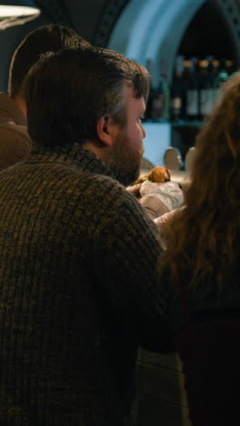Vertical-shot-of-man-eats-burger,-talks-to-wife-sitting-at-the-bar-counter-in-stylish-crowded-pub.-Loving-couple-having-dinner-on-date,-spending-weekend-together-in-restaurant.-Lifestyle-and-leisure-concept.