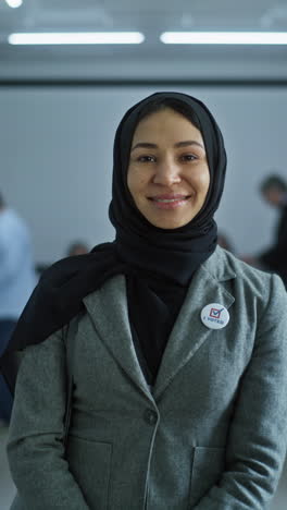 Woman-stands-in-a-modern-polling-station,-poses,-smiles-and-looks-at-camera.-Portrait-of-Muslim-woman,-United-States-of-America-elections-voter.-Background-with-voting-booths.-Concept-of-civic-duty.