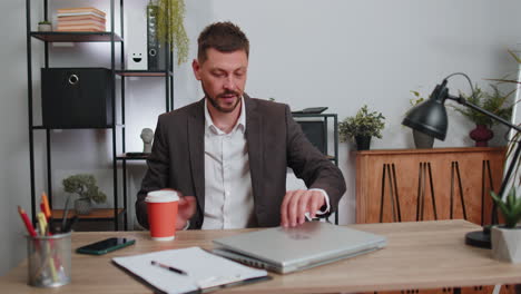 Businessman-enters-office-start-working-on-laptop-computer-at-desk-and-drinking-morning-coffee