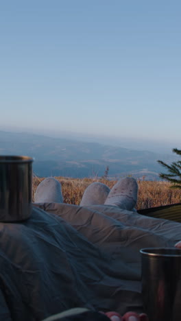 Romantic-traveler-couple-lie-in-tent-on-mountain-hill-and-drink-tea.-Two-tourists-rest-under-blanket-during-adventure-vacation.-Backpacker-family-warm-up-at-cold-windy-weather-and-admire-the-scenery.