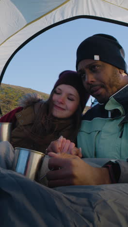 Multiethnic-backpacker-couple-sit-in-tent-on-mountain-hill:-They-hug-and-drink-tea.-Two-travelers-stopped-to-rest-during-adventure-vacation.-Romantic-hiker-family-admire-the-nature.-Vertical-shot