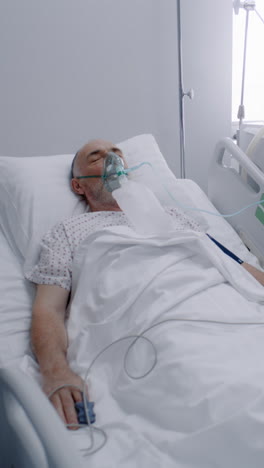 Sick-elderly-man-in-oxygen-mask-sleeps-in-bed-in-hospital-ward.-Old-patient-during-lung-ventilation.-Equipped-emergency-room-in-clinic.-Intensive-care-coronavirus-department-in-modern-medical-center.