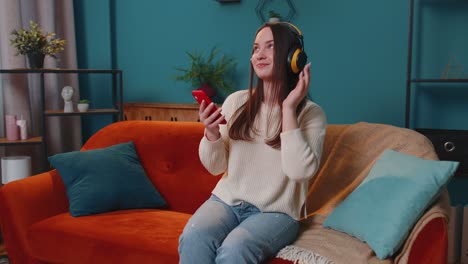 Overjoyed-young-woman-in-wireless-headphones-dancing,-singing-on-cozy-couch-in-living-room-at-home