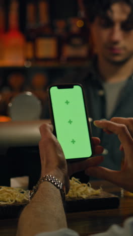 Vertical-shot-of-Mature-sports-fan-uses-mobile-phone-with-green-screen,-bets-on-football-match-online,-talks-with-bartender-and-friend.-Man-checks-bookmaker-ratings-in-app-sitting-at-bar-counter-in-pub.