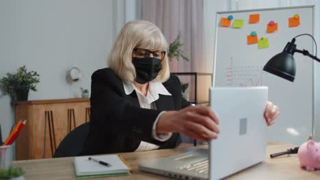 Senior-employee-wears-face-mask-works-alone-from-home-office-sits-at-desk-using-laptop-computer
