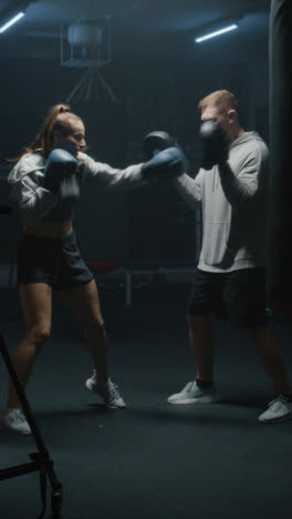 Vertical-shot-of-Female-boxer-in-boxing-gloves-hits-punching-mitts-and-practices-fighting-techniques-with-coach-in-dark-boxing-gym.-Athletic-woman-exercises-and-prepares-for-match.