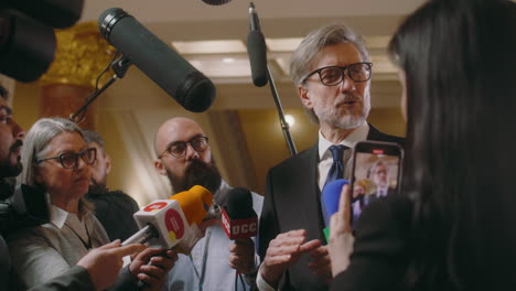 Politician-answers-press-questions-and-gives-interview-for-television-news.-Mature-diplomat-surrounded-by-crowd-of-journalists-in-parliament-hall.-Press-conference.-Inspirational-speech.-Slow-motion.
