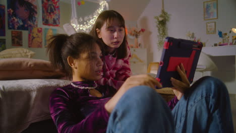 African-American-girl-sits-at-floor,-surfs-internet-using-tablet.-Asian-teen-lies-on-bed-and-watches-content-with-friend.-Happy-diverse-girls-together-spend-leisure-time-at-home.-Friends-relationship.