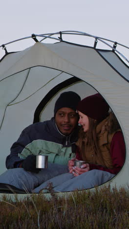 Multiethnic-couple-of-tourists-rest-on-hill-and-talk.-Young-family-sits-in-tent,-drinks-tea-and-looks-at-sunset.-African-American-man-with-Caucasian-woman-enjoys-view-in-mountains-on-their-vacation.