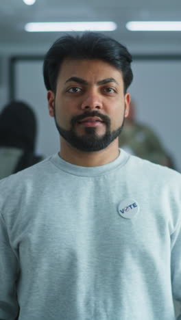 Portrait-of-Indian-man,-United-States-of-America-elections-voter.-Man-stands-in-a-modern-polling-station,-poses-and-looks-at-camera.-Background-with-voting-booths.-Civic-duty-and-patriotism-concept.