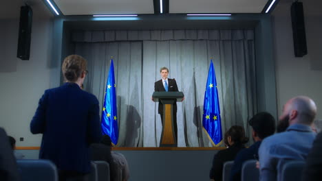 Confident-politician-makes-an-announcement,-answers-media-questions-and-gives-interview.-Mature-representative-of-the-European-Union-during-performance-at-press-conference.-Backdrop-with-EU-flags.