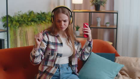 Overjoyed-young-woman-in-wireless-headphones-dancing,-singing-on-cozy-couch-in-living-room-at-home