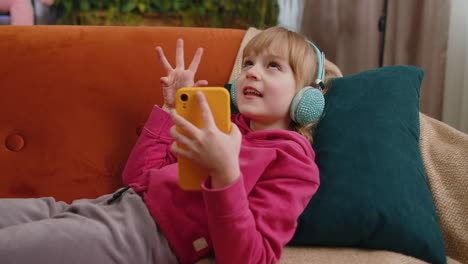 Relaxed-child-kid-girl-in-headphones-listening-energetic-disco-music-in-smartphone-lying-on-sofa
