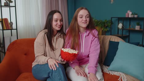 Two-girls-friends-siblings-watching-favorite-tv-show-movie,-eating-popcorn,-sitting-on-sofa-at-home
