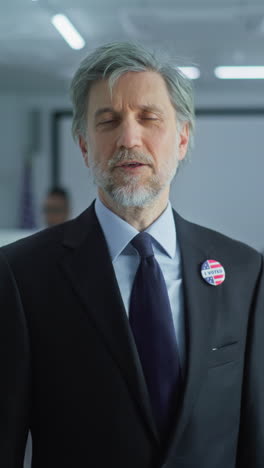 Businessman-with-badge-speaks-on-camera-and-calls-for-voting-in-modern-polling-station.-Portrait-of-man,-United-States-of-America-elections-voter.-Background-with-voting-booths.-Concept-of-civic-duty.