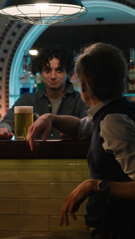 Vertical-shot-of-Young-bartender-pours-craft-beer-into-glass-and-gives-it-to-mature-man.-Male-friends-clink-glasses-and-drink-beer-sitting-at-the-bar-counter-in-stylish-pub-at-night.-Concept-of-lifestyle-and-leisure.