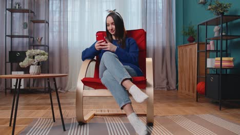 Relaxed-Caucasian-young-woman-sitting-on-home-chair-with-smartphone-scrolling,-browsing-online