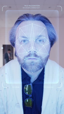Adult-man-scans-his-face-in-coworking-office.-He-touches-sensor-and-security-system-identifies-him-showing-personal-virtual-profile.-3D-hologram-of-human-biometric-facial-recognition.-Vertical-shot