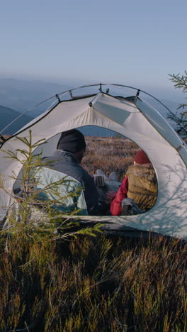 Multiethnic-tourist-couple-sit-in-tent-on-top-of-mountain-hill:-They-talk-and-drink-tea.-Two-travelers-stopped-to-rest-during-adventure-vacation.-Romantic-hiker-family-admire-the-scenery.-Vertical-shot