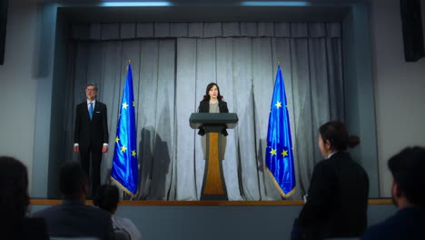 Female-politician-makes-an-announcement,-answers-journalists-questions-and-gives-interview-for-media.-Confident-representative-of-the-European-Union-during-press-conference.-Backdrop-with-EU-flags.
