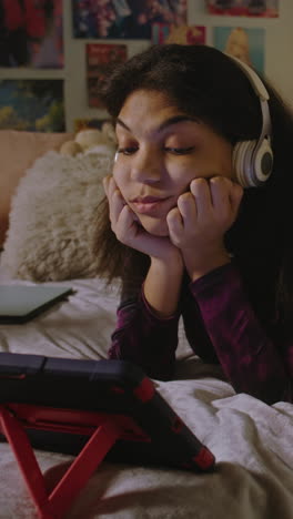 African-American-girl-looks-at-tablet-display-while-watches-movie-or-video-content.-Teenage-girl-in-headphones-lies-on-bed-in-her-room-and-uses-tablet-computer.-Vertical-shot