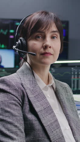 Female-trader-in-headset-sits-at-computer-with-displayed-real-time-stocks-and-looks-at-camera.-Colleagues-analyze-exchange-market-charts-on-big-screens-at-background.-Investment-and-trading.-Portrait.