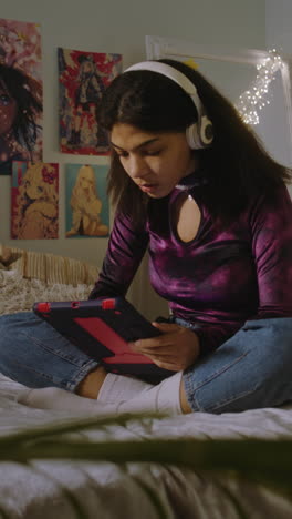 Beautiful-African-American-girl-in-headphones-sits-on-bed-in-her-room-and-surfs-internet-using-tablet.-Happy-teenager-chats-with-friends-or-listens-music.-Vertical-shot