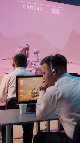 Young-female-employee-in-headset-typing-on-computer-keyboard-and-watching-video-from-Mars-research-vehicle-while-sitting-at-desk-near-colleagues-in-control-room.-Vertical-shot