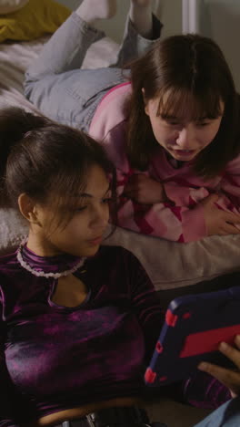 Vertical-shot-of-girl-sits-at-floor,-surfs-internet-using-tablet.-Mongolian-teen-lies-on-bed,-watches-content-with-friend.-Multi-ethnic-girls-together-spend-leisure-time-at-home.-Friends-relationship.