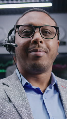 African-American-trader-in-headset-sits-at-workplace,-smiles-and-looks-into-camera.-Coworkers-analyze-exchange-market-charts-on-big-screens-at-background.-Trading-and-investment.-Dolly-shot.-Portrait.