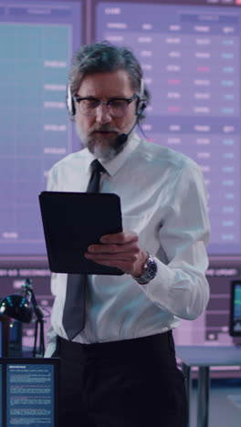 Middle-aged-bearded-man-with-headset-using-tablet-and-talking-while-working-amidst-team-of-professional-traders-in-stock-exchange-office.-Vertical-shot