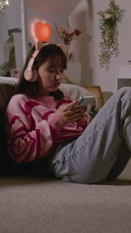 Asian-teen-girl-in-headphones-talks-by-video-chat,-surfs-the-Internet-using-mobile-phone-while-lying-on-the-bed-in-cozy-bedroom.-Girl-spending-leisure-time-and-having-fun-at-home.-Vertical-shot