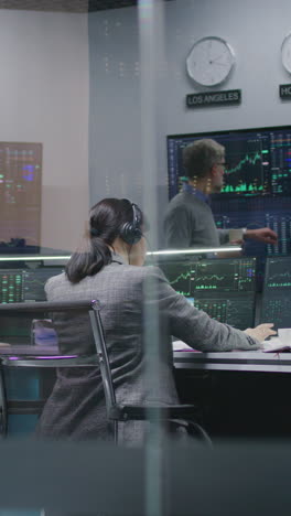 Female-financial-analyst-and-investment-manager-discuss-real-time-stocks-data-on-computer.-Colleagues-work-in-bank-office.-Big-digital-screens-showing-exchange-market-charts.-Vertical-shot