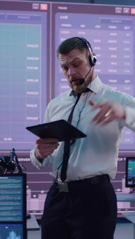 Vertical-shot-of-bearded-nervous-man-with-headset-and-tablet-adjusting-tie-and-giving-instruction-to-team-of-traders-in-stock-exchange-office