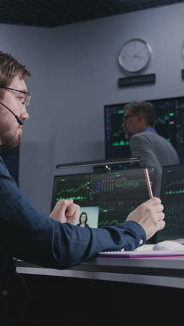 Trader-with-pen-in-hand-works-at-computer-with-displayed-real-time-stocks.-Colleagues-analyze-exchange-market-charts-on-big-screens-at-background.-Office-illuminated-by-blue-light.-Vertical-shot.