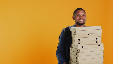 Male-pizzeria-courier-carrying-huge-pile-of-pizza-boxes-in-studio