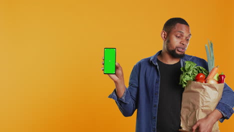 Young-guy-with-a-bag-of-fresh-groceries-showing-greenscreen-on-his-smartphone