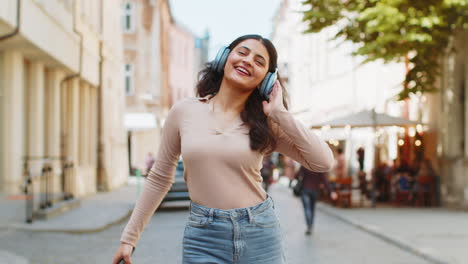 Happy-Indian-woman-in-wireless-headphones-listening-music-in-smartphone-app-dancing-on-city-street