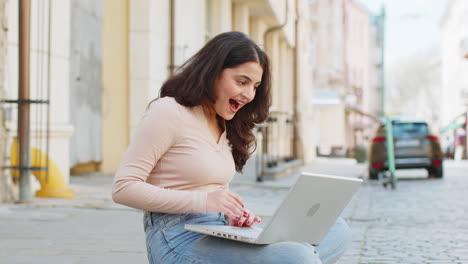 Indian-happy-woman-working-on-laptop-celebrate-success-win-money-sitting-on-urban-street-in-city