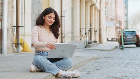 Woman-freelancer-working-online-distant-job-with-laptop-sitting-on-city-street-browsing-website